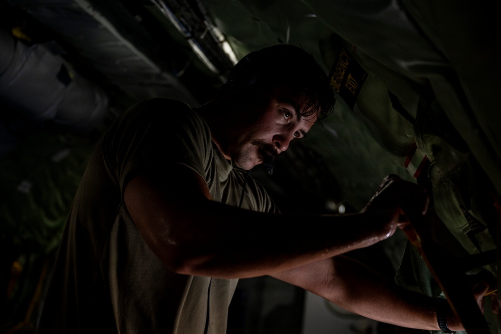 A man wearing an olive/military green t-shirt is working inside what appears to be a military aircraft, likely performing maintenance or inspection tasks. The environment is dimly lit, with the interior walls showing green military-style padding and a visible 'EMERGENCY EXIT' label. The dramatic low-key lighting highlights the man's focused expression as he manipulates equipment or panels.