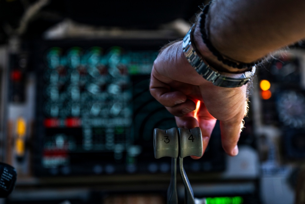 A person's hand, wearing a metal-band wristwatch, is gripping what appears to be throttle levers numbered '3' and '4' in an aircraft cockpit. The background shows a complex instrument panel with illuminated displays, red indicators, and various controls consistent with a large military or commercial aircraft flight deck. The image is a close-up shot with shallow depth of field, keeping the throttle levers and hand in sharp focus.