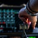 A person's hand, wearing a metal-band wristwatch, is gripping what appears to be throttle levers numbered '3' and '4' in an aircraft cockpit. The background shows a complex instrument panel with illuminated displays, red indicators, and various controls consistent with a large military or commercial aircraft flight deck. The image is a close-up shot with shallow depth of field, keeping the throttle levers and hand in sharp focus.