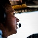 A close-up profile shot of a pilot seated in an aircraft cockpit, wearing aviation headset with boom microphone. The image is dramatically lit with strong contrast between shadow and light, capturing the pilot gazing forward through the cockpit windshield. Overhead cockpit panels with switches and yellow markings are partially visible in the background.
