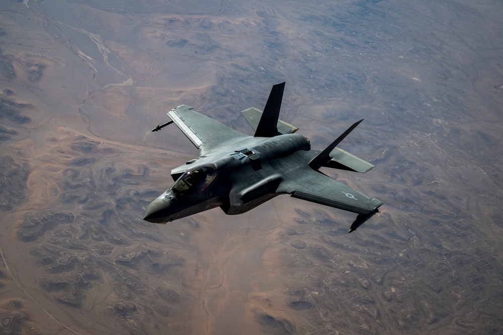 A single-seat stealth fighter jet, identifiable as an F-35 Lightning II, is photographed from above in flight over an arid, desert terrain. The aircraft is viewed from a slightly elevated angle, revealing its top surface, cockpit, and delta-wing configuration. The terrain below appears to be a dry, barren landscape with visible erosion patterns and riverbeds.