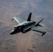 A single-seat stealth fighter jet, identifiable as an F-35 Lightning II, is photographed from above in flight over an arid, desert terrain. The aircraft is viewed from a slightly elevated angle, revealing its top surface, cockpit, and delta-wing configuration. The terrain below appears to be a dry, barren landscape with visible erosion patterns and riverbeds.