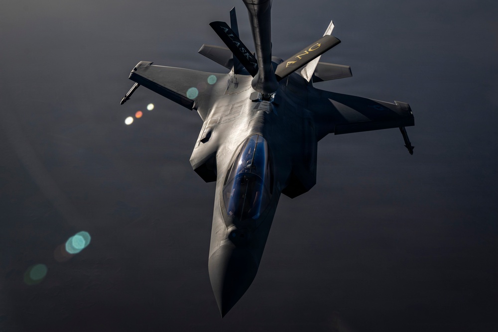 An F-22 Raptor stealth fighter jet is captured from above during an aerial refueling operation at night or in low-light conditions. The aircraft bears markings reading 'ALASKA' and 'ANG' (Alaska Air National Guard) on its tail fins. A refueling boom is visible connecting from above to the aircraft's receptacle.