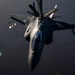 An F-22 Raptor stealth fighter jet is captured from above during an aerial refueling operation at night or in low-light conditions. The aircraft bears markings reading 'ALASKA' and 'ANG' (Alaska Air National Guard) on its tail fins. A refueling boom is visible connecting from above to the aircraft's receptacle.