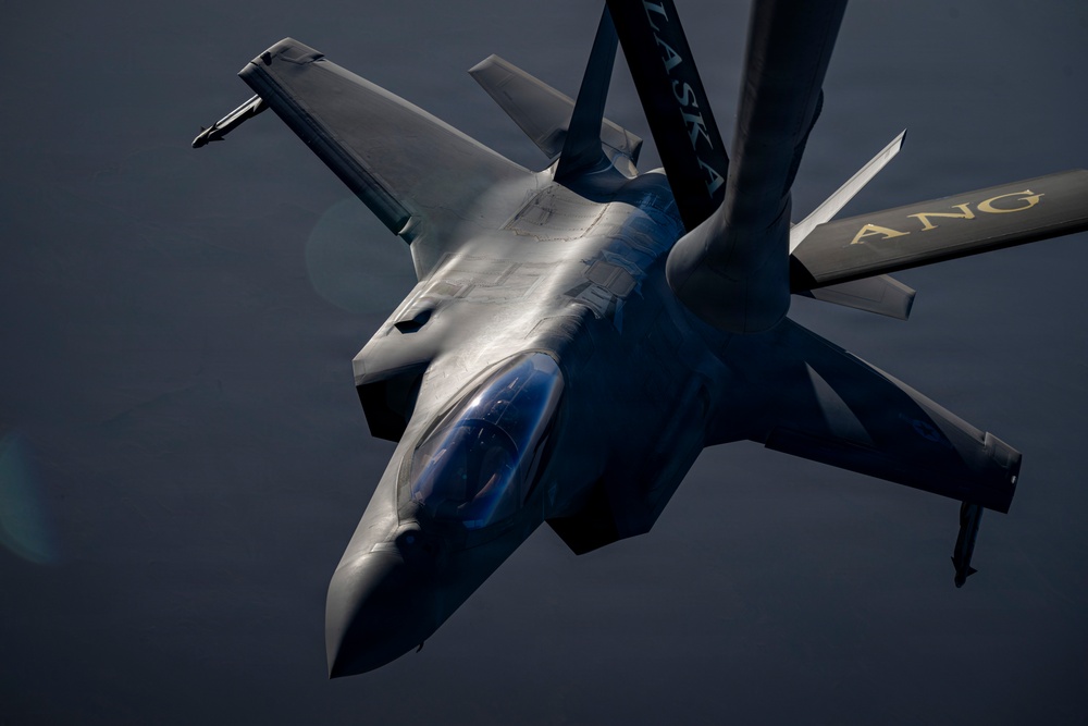 A military fighter jet, identifiable as an F-22 Raptor, is photographed in close proximity to a tanker aircraft during an aerial refueling operation. The tanker's boom and wing are visible in the upper portion of the frame, bearing the markings 'ALASKA' and 'ANG' (Air National Guard) in gold lettering on a dark blue surface. The image is taken from the tanker's perspective looking down at the receiver aircraft.