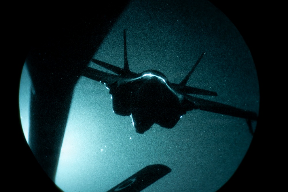 A military jet aircraft is captured in silhouette through what appears to be a night-vision or circular optical device, likely a refueling boom operator's window or periscope view. The aircraft is seen from below/behind during what appears to be an aerial refueling operation at night. The teal-green tint and circular vignette are characteristic of night-vision optics or a specialized observation window.