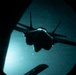 A military jet aircraft is captured in silhouette through what appears to be a night-vision or circular optical device, likely a refueling boom operator's window or periscope view. The aircraft is seen from below/behind during what appears to be an aerial refueling operation at night. The teal-green tint and circular vignette are characteristic of night-vision optics or a specialized observation window.