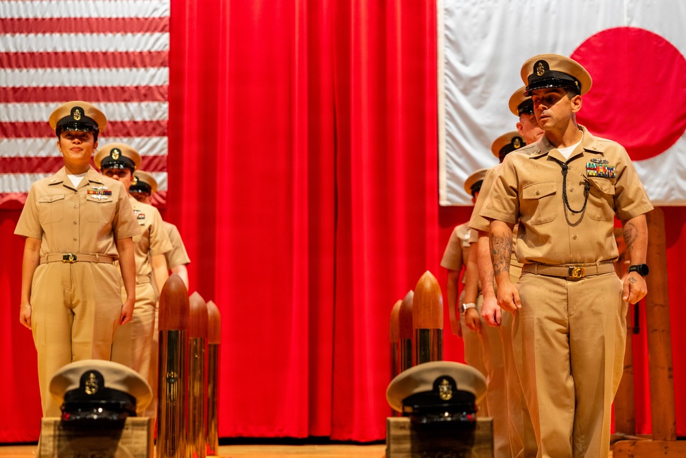 A formal U.S. Navy Chief Petty Officer pinning or advancement ceremony is taking place on a stage. Multiple sailors in tan/khaki Service Dress Khaki uniforms stand at attention before a red curtain backdrop flanked by the American flag and the Japanese flag, indicating the ceremony is likely held at a U.S. military installation in Japan. Navy covers (hats) and ceremonial shell casings are placed on wooden pedestals in the foreground.