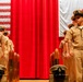 A formal U.S. Navy Chief Petty Officer pinning or advancement ceremony is taking place on a stage. Multiple sailors in tan/khaki Service Dress Khaki uniforms stand at attention before a red curtain backdrop flanked by the American flag and the Japanese flag, indicating the ceremony is likely held at a U.S. military installation in Japan. Navy covers (hats) and ceremonial shell casings are placed on wooden pedestals in the foreground.