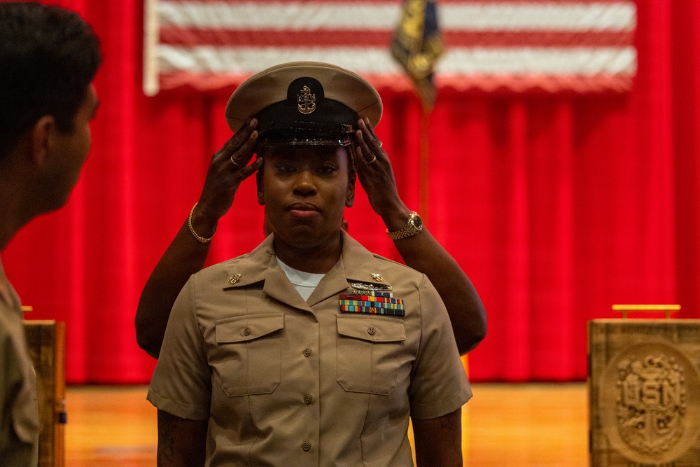A U.S. Navy service member is having a chief petty officer cover (combination cap) placed on their head during what appears to be a Chief Petty Officer pinning or frocking ceremony. The individual is wearing the tan/khaki Navy Working Uniform with multiple service ribbons and insignia. The ceremony is taking place in a formal venue decorated with an American flag and red curtains, with a USN-emblazoned podium visible.
