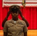 A U.S. Navy service member is having a chief petty officer cover (combination cap) placed on their head during what appears to be a Chief Petty Officer pinning or frocking ceremony. The individual is wearing the tan/khaki Navy Working Uniform with multiple service ribbons and insignia. The ceremony is taking place in a formal venue decorated with an American flag and red curtains, with a USN-emblazoned podium visible.