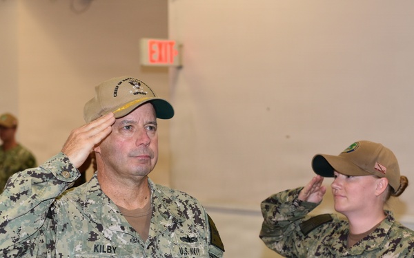 A group of U.S. Navy personnel in NWU Type III (digital camouflage) uniforms are rendering a salute in what appears to be an indoor military facility. The central figure, identified by name tape as 'KILBY,' wears rank insignia consistent with a senior officer and a cap with 'Chief of Naval Operations' text. Multiple sailors are visible saluting in formation, suggesting a formal military ceremony or official visit.
