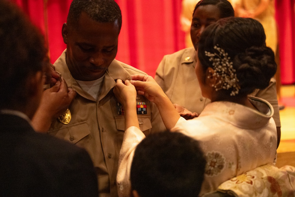 A military promotion or pinning ceremony is taking place, with a service member in tan/khaki U.S. Navy or military uniform receiving new insignia pinned onto his collar or chest. A woman in formal attire with an ornate hair accessory is performing the pinning, while others assist on the opposite side. The red curtain backdrop and formal dress of attendees suggest an official ceremonial setting.