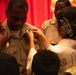 A military promotion or pinning ceremony is taking place, with a service member in tan/khaki U.S. Navy or military uniform receiving new insignia pinned onto his collar or chest. A woman in formal attire with an ornate hair accessory is performing the pinning, while others assist on the opposite side. The red curtain backdrop and formal dress of attendees suggest an official ceremonial setting.