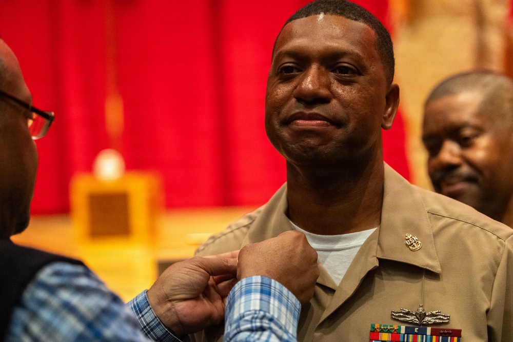 A U.S. Navy sailor in tan service uniform is having an insignia or device pinned onto his uniform during what appears to be a formal military ceremony. The individual displays an emotional expression, suggesting this is a significant milestone such as a promotion or award ceremony. The setting appears to be an indoor ceremonial hall with red curtains in the background.