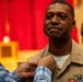 A U.S. Navy sailor in tan service uniform is having an insignia or device pinned onto his uniform during what appears to be a formal military ceremony. The individual displays an emotional expression, suggesting this is a significant milestone such as a promotion or award ceremony. The setting appears to be an indoor ceremonial hall with red curtains in the background.