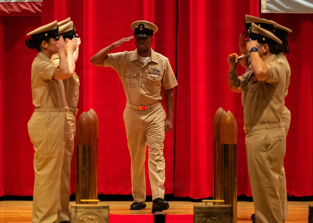 Three U.S. Navy personnel in khaki service uniforms are on a stage with a red curtain backdrop, performing a formal salute ceremony. The central figure, a senior enlisted sailor wearing a combination cover and displaying ribbons and a warfare device, is returning salutes from two flanking sailors. The setting appears to be an official military ceremony, likely a change of command, retirement, or advancement event.