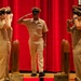 Three U.S. Navy personnel in khaki service uniforms are on a stage with a red curtain backdrop, performing a formal salute ceremony. The central figure, a senior enlisted sailor wearing a combination cover and displaying ribbons and a warfare device, is returning salutes from two flanking sailors. The setting appears to be an official military ceremony, likely a change of command, retirement, or advancement event.