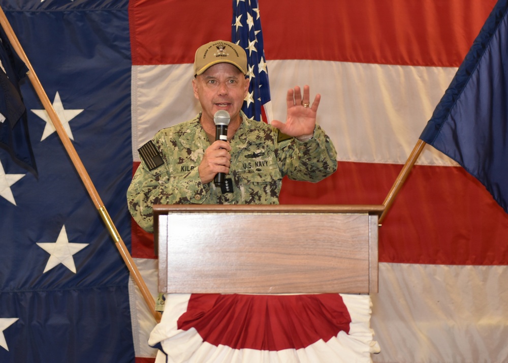 A U.S. Navy officer, identified by the name tape 'KILB' (partially visible), is speaking at a podium while holding a microphone and raising one hand. He is wearing Navy Working Uniform (NWU) camouflage and a tan cap with military insignia. The setting appears to be a formal military event or ceremony, decorated with large American flags and patriotic bunting.