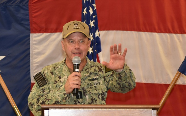 A U.S. Navy officer, identified by the name tape 'KILB' (partially visible), is speaking at a podium while holding a microphone and raising one hand. He is wearing Navy Working Uniform (NWU) camouflage and a tan cap with military insignia. The setting appears to be a formal military event or ceremony, decorated with large American flags and patriotic bunting.