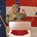 A U.S. Navy officer, identified by the name tape 'KILB' (partially visible), is speaking at a podium while holding a microphone and raising one hand. He is wearing Navy Working Uniform (NWU) camouflage and a tan cap with military insignia. The setting appears to be a formal military event or ceremony, decorated with large American flags and patriotic bunting.