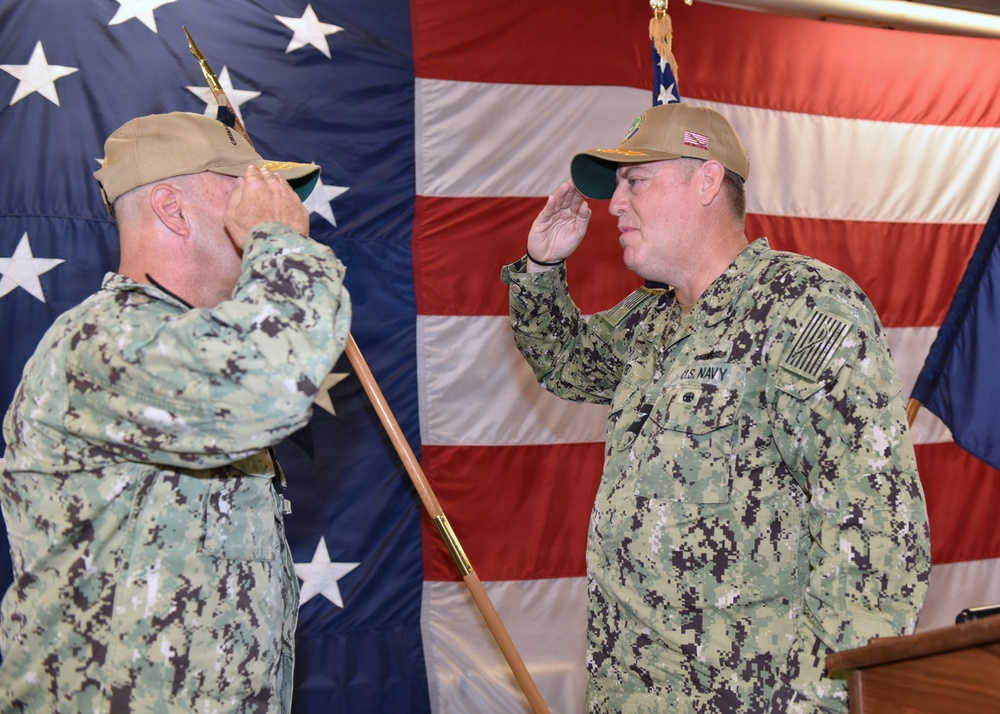 Two U.S. Navy personnel in Navy Working Uniform (NWU) Type III camouflage are exchanging salutes during what appears to be a formal military ceremony. Both individuals are wearing tan/coyote brown caps and are positioned in front of a large American flag backdrop. The setting suggests a change of command or similar official military ceremony.