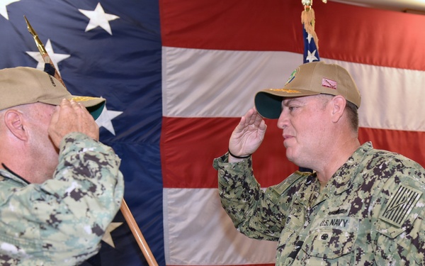 Two U.S. Navy personnel in Navy Working Uniform (NWU) Type III camouflage are exchanging salutes during what appears to be a formal military ceremony. Both individuals are wearing tan/coyote brown caps and are positioned in front of a large American flag backdrop. The setting suggests a change of command or similar official military ceremony.