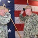 Two U.S. Navy personnel in Navy Working Uniform (NWU) Type III camouflage are exchanging salutes during what appears to be a formal military ceremony. Both individuals are wearing tan/coyote brown caps and are positioned in front of a large American flag backdrop. The setting suggests a change of command or similar official military ceremony.