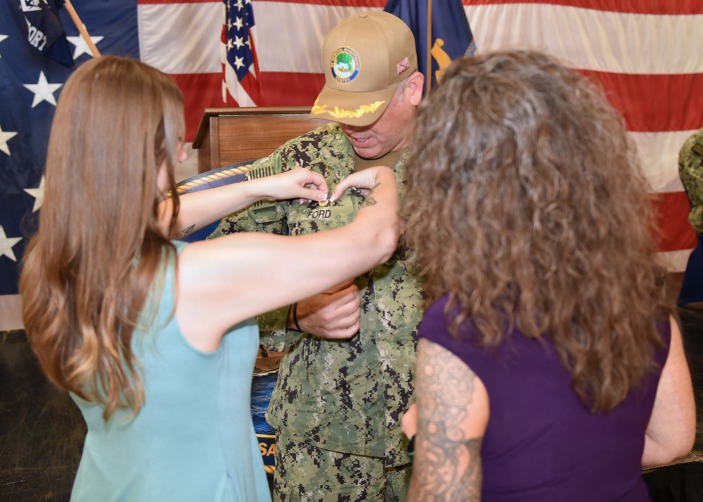 A military promotion or pinning ceremony is taking place, with a service member in U.S. Navy digital camouflage (NWU Type III) uniform receiving a pin or insignia on his chest. Two women, one in a teal dress and one in a purple top, are participating in the pinning. An American flag and what appears to be a unit or command flag are visible in the background.
