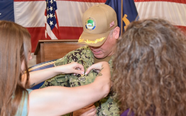 A military promotion or pinning ceremony is taking place, with a service member in U.S. Navy digital camouflage (NWU Type III) uniform receiving a pin or insignia on his chest. Two women, one in a teal dress and one in a purple top, are participating in the pinning. An American flag and what appears to be a unit or command flag are visible in the background.
