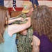 A military promotion or pinning ceremony is taking place, with a service member in U.S. Navy digital camouflage (NWU Type III) uniform receiving a pin or insignia on his chest. Two women, one in a teal dress and one in a purple top, are participating in the pinning. An American flag and what appears to be a unit or command flag are visible in the background.