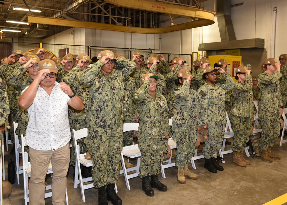 A group of U.S. Navy personnel in NWU Type III (woodland camouflage) uniforms are simultaneously placing or adjusting matching tan/brown caps during what appears to be a formal ceremony or event held inside an industrial or hangar-type facility. One civilian male in a white floral shirt and khaki pants participates alongside the uniformed personnel. White folding chairs are arranged in rows behind the group, suggesting a seated ceremony.