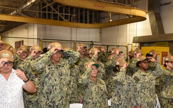 A group of U.S. Navy personnel in NWU Type III (woodland camouflage) uniforms are simultaneously placing or adjusting matching tan/brown caps during what appears to be a formal ceremony or event held inside an industrial or hangar-type facility. One civilian male in a white floral shirt and khaki pants participates alongside the uniformed personnel. White folding chairs are arranged in rows behind the group, suggesting a seated ceremony.