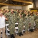 A group of U.S. Navy personnel in NWU Type III (woodland camouflage) uniforms are simultaneously placing or adjusting matching tan/brown caps during what appears to be a formal ceremony or event held inside an industrial or hangar-type facility. One civilian male in a white floral shirt and khaki pants participates alongside the uniformed personnel. White folding chairs are arranged in rows behind the group, suggesting a seated ceremony.