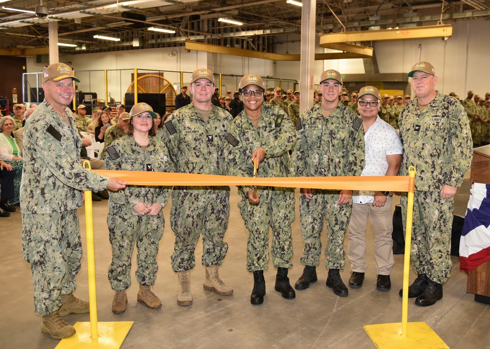 A group of U.S. Navy personnel in camouflage uniforms (NWU Type III) pose for a ribbon-cutting ceremony inside what appears to be a large industrial or warehouse facility. Seven individuals stand behind an orange safety barrier/ribbon stretched between two yellow stanchions, with one civilian present among the uniformed members. A large crowd of military personnel is visible in the background.