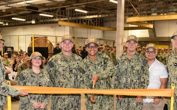 A group of U.S. Navy personnel in camouflage uniforms (NWU Type III) pose for a ribbon-cutting ceremony inside what appears to be a large industrial or warehouse facility. Seven individuals stand behind an orange safety barrier/ribbon stretched between two yellow stanchions, with one civilian present among the uniformed members. A large crowd of military personnel is visible in the background.