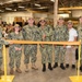 A group of U.S. Navy personnel in camouflage uniforms (NWU Type III) pose for a ribbon-cutting ceremony inside what appears to be a large industrial or warehouse facility. Seven individuals stand behind an orange safety barrier/ribbon stretched between two yellow stanchions, with one civilian present among the uniformed members. A large crowd of military personnel is visible in the background.