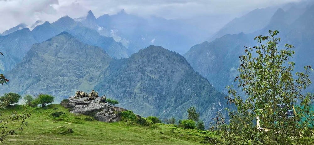 A panoramic mountain landscape featuring multiple layered ridgelines shrouded in mist and low clouds. In the foreground, a group of people are seated on a prominent rocky outcrop surrounded by lush green alpine meadows. A solar panel and a tree are visible on the right side, suggesting a remote habitation or outpost.