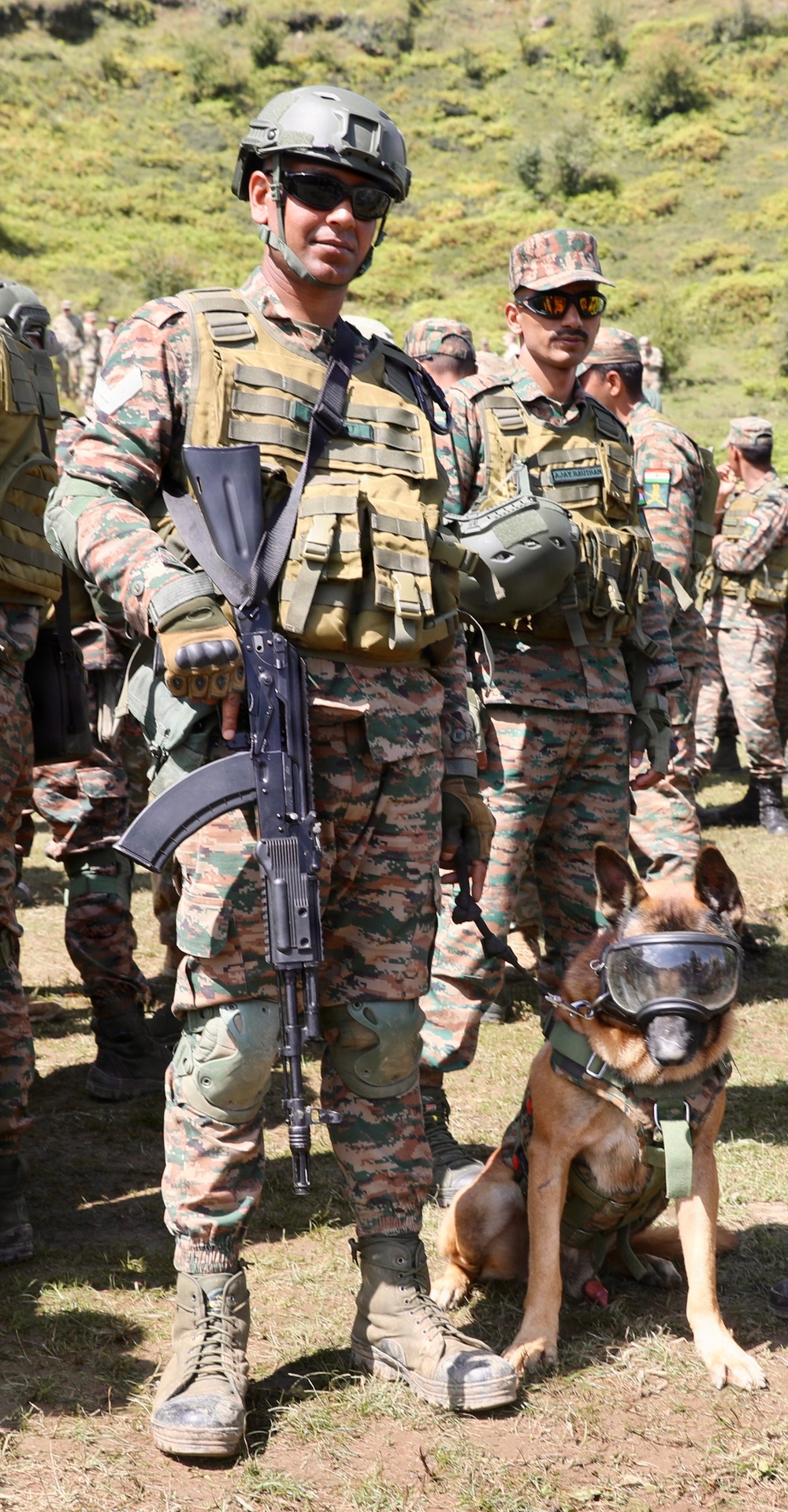 A military soldier in full combat gear stands at attention holding an assault rifle, accompanied by a military working dog wearing tactical goggles and a harness. The soldier wears digital camouflage uniform, tactical vest, ballistic helmet, and sunglasses. Multiple other soldiers in similar uniforms are visible in the background against a green hillside.