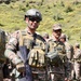 A military soldier in full combat gear stands at attention holding an assault rifle, accompanied by a military working dog wearing tactical goggles and a harness. The soldier wears digital camouflage uniform, tactical vest, ballistic helmet, and sunglasses. Multiple other soldiers in similar uniforms are visible in the background against a green hillside.