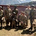 A group of military personnel from what appear to be two different armed forces pose together for a photograph in a mountainous, forested setting. The group includes approximately eight soldiers accompanied by three military working dogs in tactical gear. The setting appears to be a joint military exercise or training event, evidenced by the mixed uniforms and cooperative posture.
