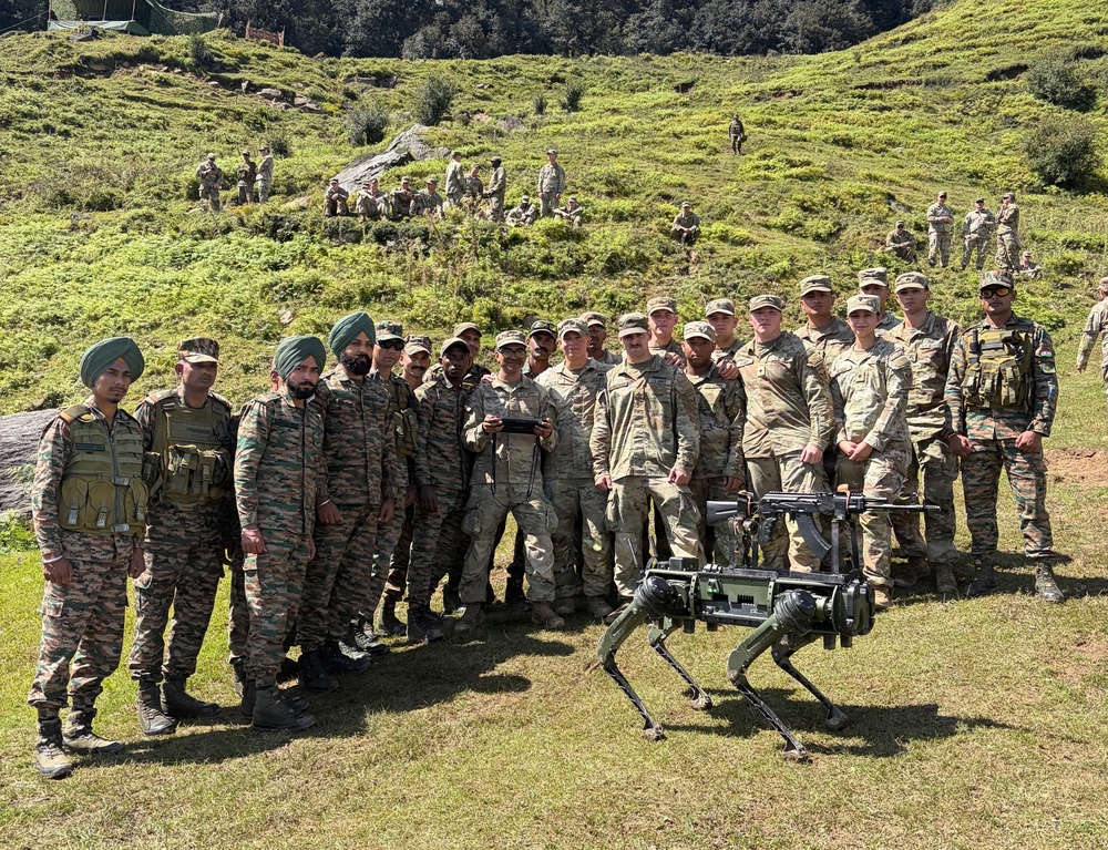 A group of military personnel from two different armed forces pose together outdoors on a grassy hillside terrain. A quadruped robotic dog, equipped with what appears to be a mounted weapon system, is positioned in the foreground. The gathering appears to be a joint military exercise or demonstration involving advanced robotic technology.