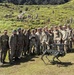 A group of military personnel from two different armed forces pose together outdoors on a grassy hillside terrain. A quadruped robotic dog, equipped with what appears to be a mounted weapon system, is positioned in the foreground. The gathering appears to be a joint military exercise or demonstration involving advanced robotic technology.