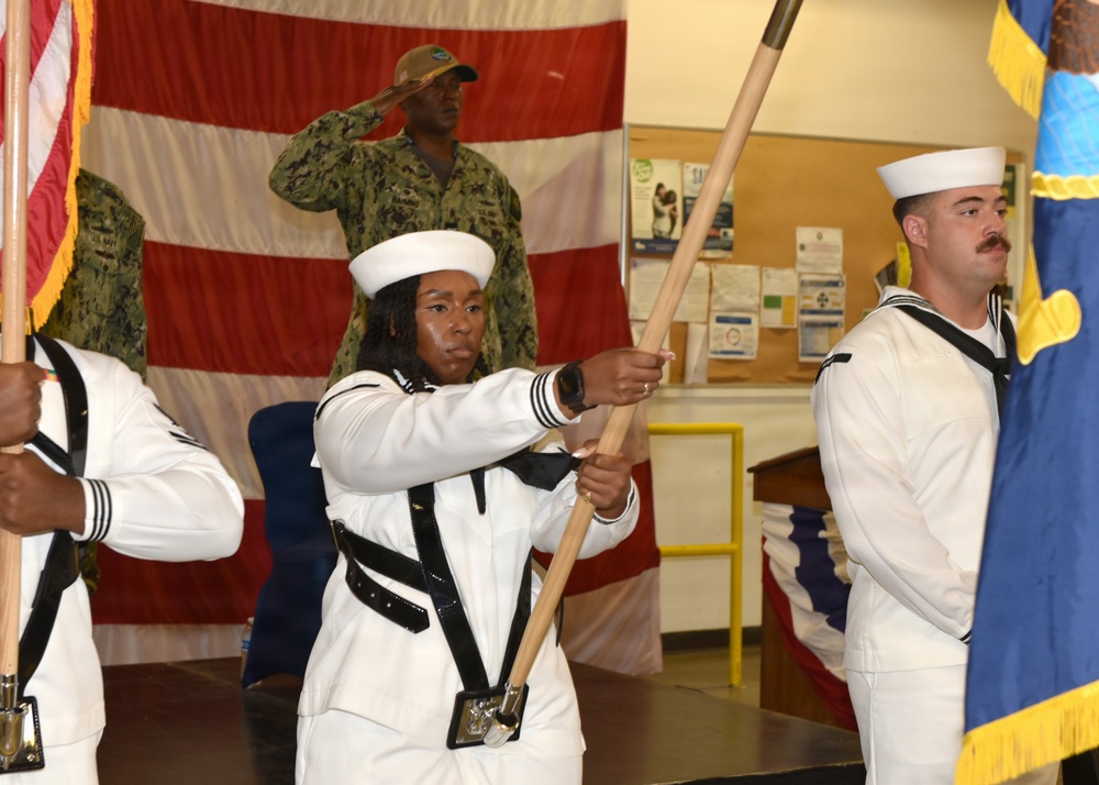 A U.S. Navy color guard ceremony is taking place indoors, with sailors in dress white uniforms carrying flags on wooden staffs. In the background, a service member in Navy Working Uniform (NWU) camouflage is rendering a salute. The setting appears to be a military facility or community center decorated with a large American flag backdrop.