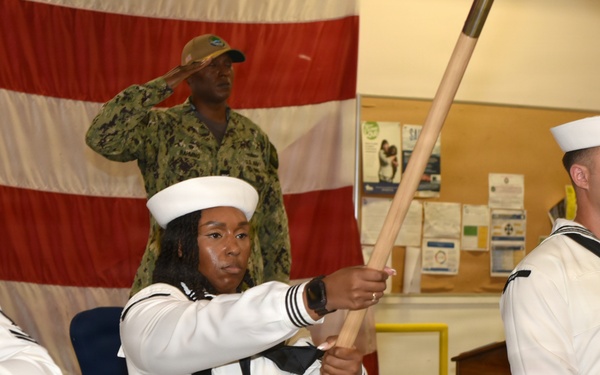 A U.S. Navy color guard ceremony is taking place indoors, with sailors in dress white uniforms carrying flags on wooden staffs. In the background, a service member in Navy Working Uniform (NWU) camouflage is rendering a salute. The setting appears to be a military facility or community center decorated with a large American flag backdrop.