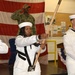 A U.S. Navy color guard ceremony is taking place indoors, with sailors in dress white uniforms carrying flags on wooden staffs. In the background, a service member in Navy Working Uniform (NWU) camouflage is rendering a salute. The setting appears to be a military facility or community center decorated with a large American flag backdrop.