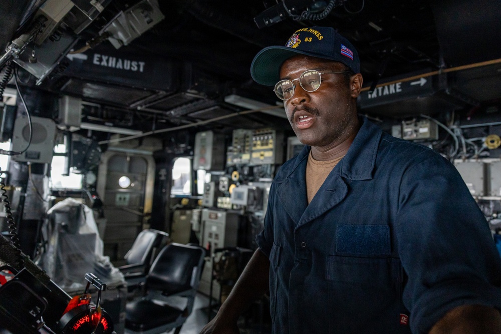 A U.S. Navy sailor stands on the bridge of a naval vessel, wearing a dark blue uniform and a ship's ball cap. The cap bears the text 'PAUL JONES 53' and an American flag patch, indicating assignment to USS Paul Jones (DDG-53). The sailor appears to be speaking or giving a briefing in the ship's pilothouse/bridge environment.