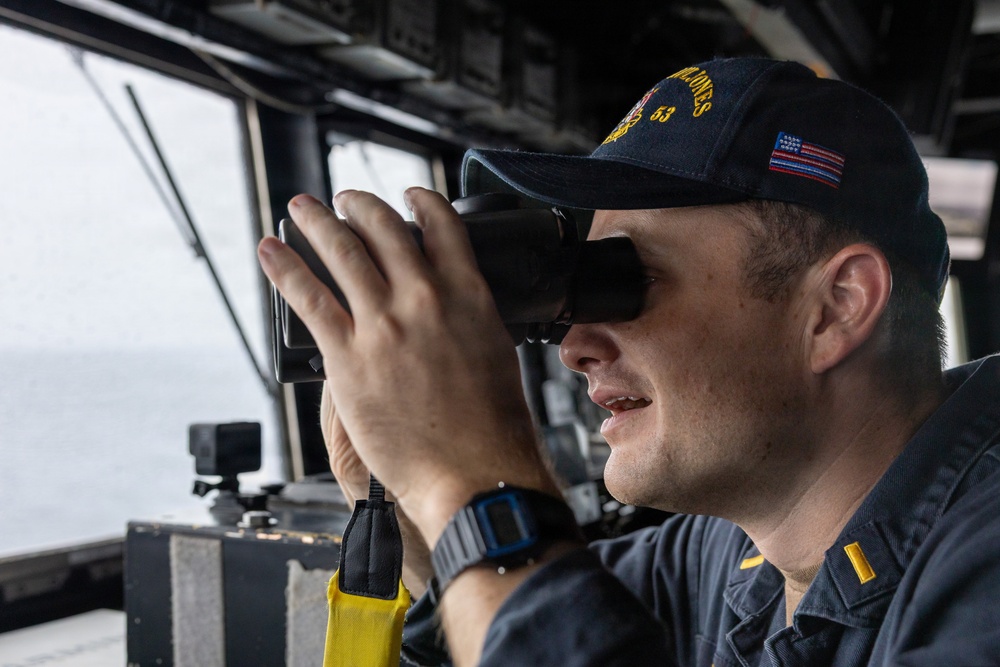 A U.S. Navy officer is using binoculars while standing watch on the bridge of a naval vessel. The officer is wearing a Navy ball cap with the number '53' visible, suggesting assignment to a specific ship. The setting appears to be an active bridge watch station with open ocean visible through the windows.