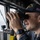 John Paul Jones Sailor Stands Watch A U.S. Navy officer is using binoculars while standing watch on the bridge of a naval vessel. The officer is wearing a Navy ball cap with the number '53' visible, suggesting assignment to a specific ship. The setting appears to be an active bridge watch station with open ocean visible through the windows.