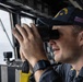 A U.S. Navy officer is using binoculars while standing watch on the bridge of a naval vessel. The officer is wearing a Navy ball cap with the number '53' visible, suggesting assignment to a specific ship. The setting appears to be an active bridge watch station with open ocean visible through the windows.
